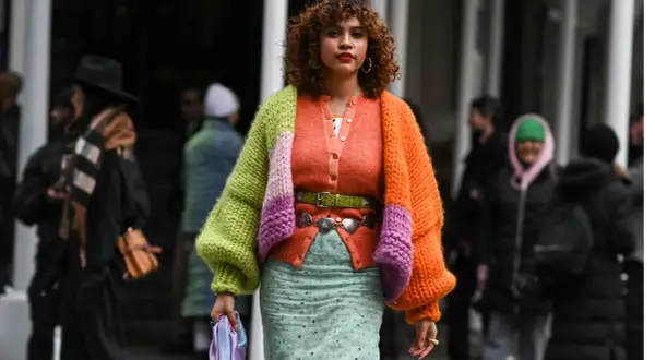 Bunte Strickjacke kombinieren: Ein Outfit mit Strickjacke in Orange, Grün und Lila, dazu ein gemusterter Rock und ein grüner Gürtel.