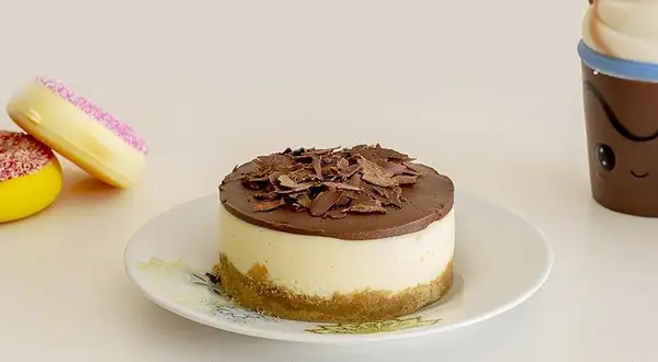 Mini cheesecake al cioccolato con base croccante e copertura di cioccolato fondente, accompagnato da due ciambelle colorate e una tazza sorridente.