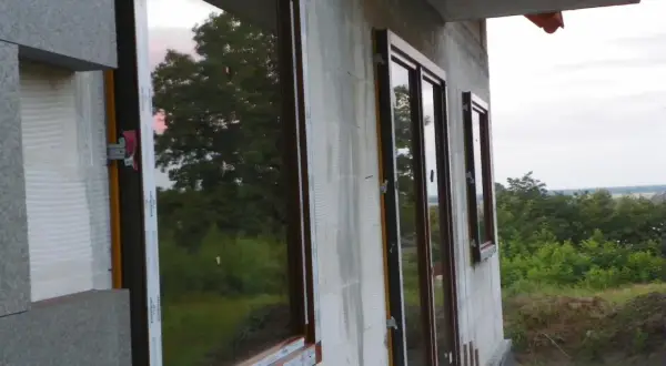 Okna z brązowymi ramami zamontowane w ścianie z bloczków. Widać fragmenty izolacji i zewnętrzny krajobraz. To przykład ciepłego montażu.