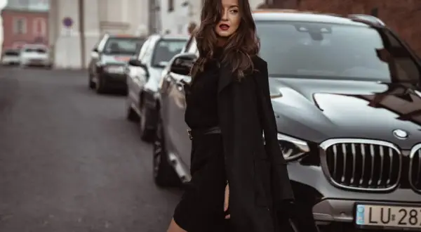 Elegancka kobieta w czarnej sukience swetrowej i płaszczu stoi obok luksusowego BMW na ulicy.