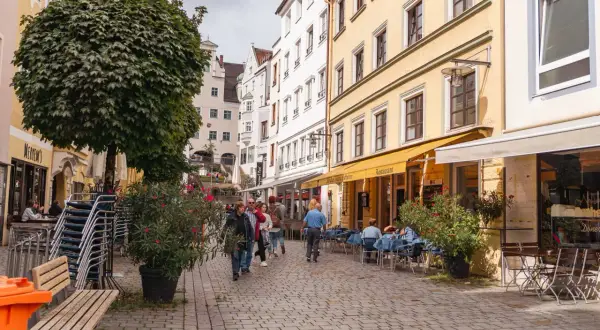 Kempten Geheimtipps: Menschen schlendern durch eine charmante Gasse mit Restaurants und Cafés, umgeben von historischen Gebäuden und üppigem Grün.