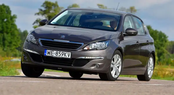 Ciemnoszary Peugeot 308 2015 z polskimi tablicami WE 859KV jedzie po drodze wśród zieleni.