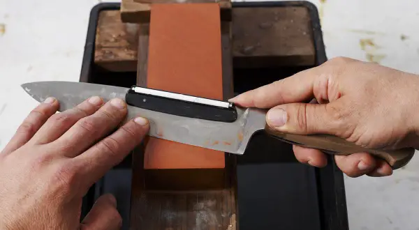Händer slipar en kniv mot en orange sten. En vass kniv är viktig för matlagning.
