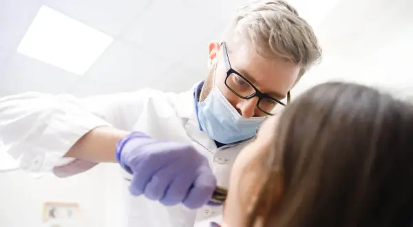 Dentysta w masce i rękawiczkach wykonuje borowanie zęba pacjentowi.