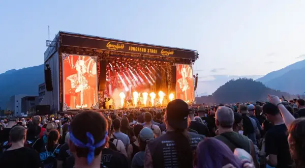 A massive crowd enjoys a performance at the Greenfield festival, with a large screen displaying a performer and flames erupting on stage.