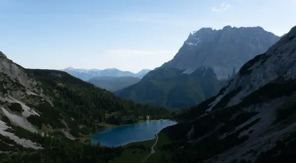 Ein Bergsee liegt eingebettet zwischen steilen Hängen, die wie ein vorderer Drachenkopf wirken. Nadelbäume bedecken die Flanken, im Hintergrund ragen weitere Gipfel auf.