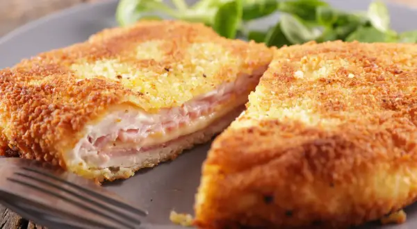 Cotoletta alla valdostana dorata e croccante, farcita con prosciutto e formaggio filante, servita su un piatto grigio con un contorno di insalata verde.