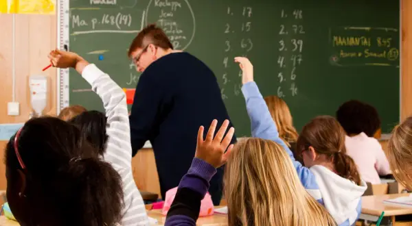 Schüler heben in einer finnischen Schule die Hände. Eine Lehrerin steht vor einer Tafel mit mathematischen Aufgaben.