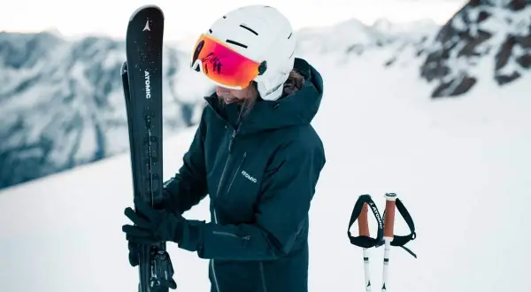 Femme en tenue de ski, tenant ses skis, portant un casque de ski avec visière intégrée.
