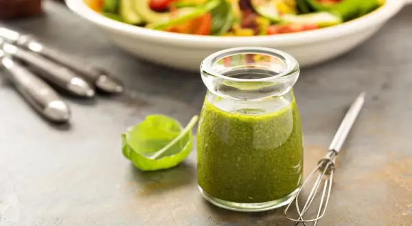 Grünes Dressing in einem Glas mit Salat und Kräutern im Hintergrund.