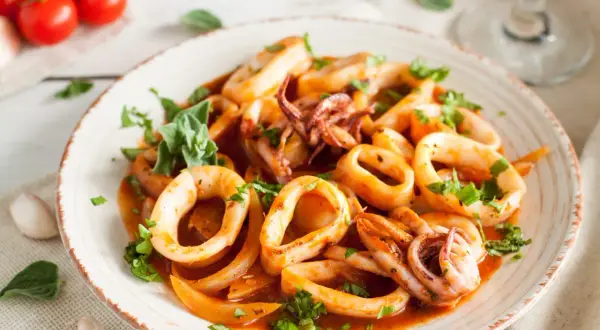 Délicieuse recette encornet à la sauce tomate, garnie de persil frais. Des tomates cerises et de l'ail sont disposés sur le côté.