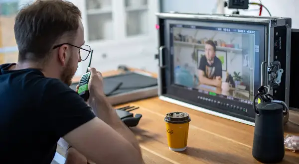 Producenci na planie filmowym czuwają nad realizacją, jeden z nich rozmawia przez krótkofalówkę, monitorując obraz na ekranie.