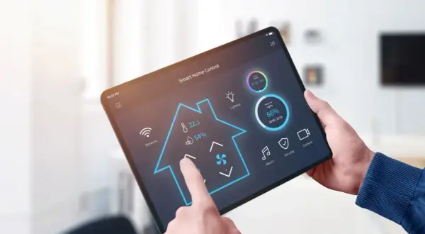 Une main utilise une tablette pour contrôler une application domotique gratuite. L'interface affiche la température, la lumière et d'autres fonctions de la maison.