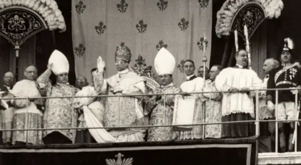 Uroczysta procesja z papieżem w tiarze, jego nakrycie głowy jest bogato zdobione. W tle inni duchowni i wierni.