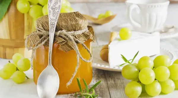 Un vasetto di confettura di uva bianca, con un cucchiaio d'argento, uva fresca, formaggio e una tazza.