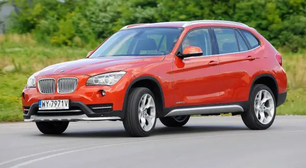 Pomarańczowe BMW E84 X1 z polskimi tablicami rejestracyjnymi jedzie po drodze, otoczone zielenią.