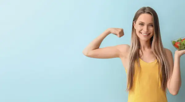 Donna sorridente mostra il bicipite e una ciotola di insalata, simbolo di una dieta per stitichezza menù settimanale salutare e benessere.