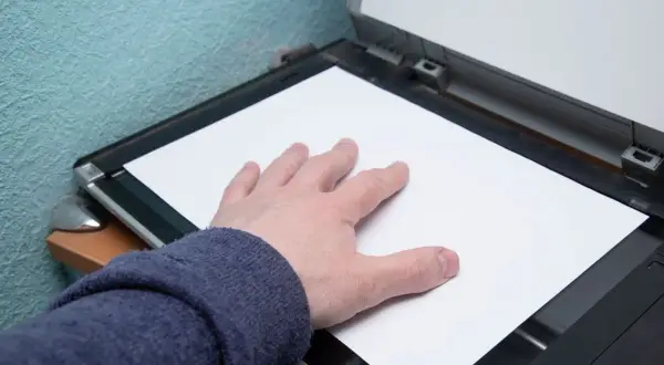 Dłoń dociska białą kartkę papieru do otwartego skanera.