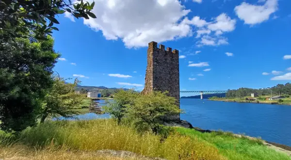 Torre medieval junto a un río azul en Catoira, Galicia. Un puente cruza el agua al fondo.