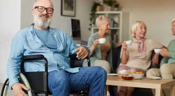 Uomo sorridente in carrozzina, con amici in sottofondo. La sua storia di invalidità civile patologie riconosciute è un esempio di resilienza.
