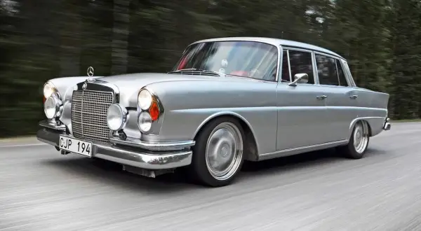 Silver Mercedes från 60-talet glider fram på en väg, omgiven av skog. Klassiska bilar som denna är tidlösa.
