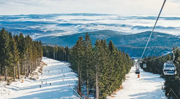 Skifahrer genießen den Winter in Tschechien. Eine Seilbahn schwebt über schneebedeckte Hänge, umgeben von dichten Fichtenwäldern und einer nebligen Berglandschaft.