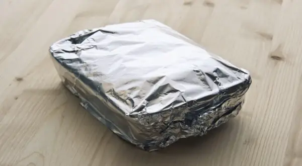 Ein rechteckiges Paket, in glänzende Alufolie gewickelt, liegt auf einem Holztisch. Die Frage, welche Seite der Alufolie giftig ist, bleibt unbeantwortet.