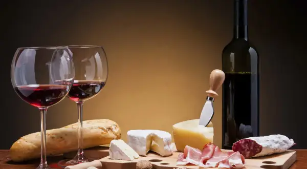 Deux verres de vin rouge, une bouteille, du pain, des fromages, de la charcuterie et des noix. Un accord parfait pour un vin pour fromage et charcuterie.