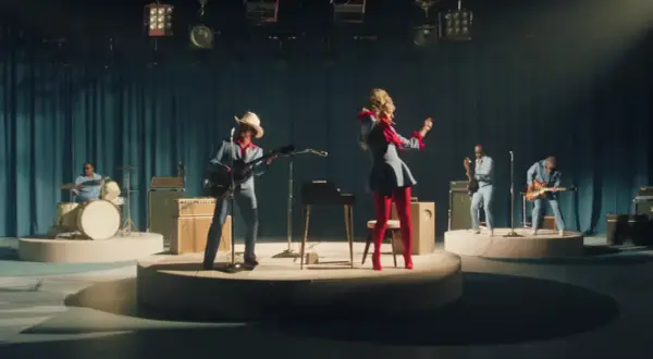 Lady Gaga w czerwonych rajstopach i niebieskiej sukience tańczy na scenie, podczas gdy zespół gra. Tekst "die with a smile" może być inspiracją do tej sceny.