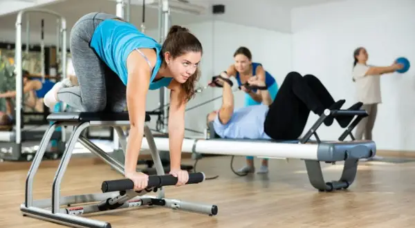Frauen trainieren im Studio. Eine macht eine Übung auf einem Gerät, während andere im Hintergrund an Pilates-Geräten arbeiten. Das ist Zirkeltraining.