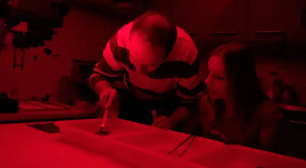 W ciemnym pokoju, przy czerwonym świetle, mężczyzna i dziewczyna pracują przy wywoływaniu zdjęć.
