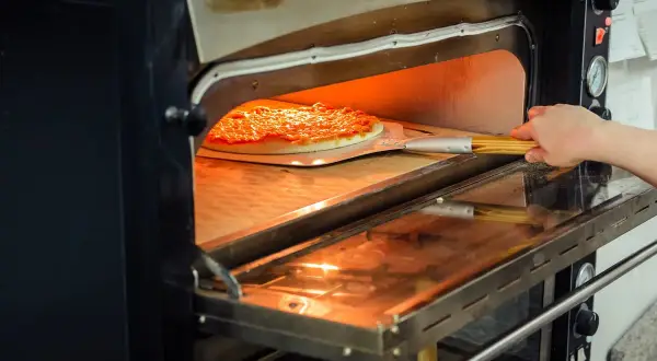 Une main glisse une pizza dans un four à pizza professionnel. La chaleur intense du four est visible, promettant une cuisson parfaite.