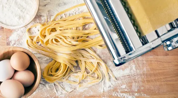 Frischer Tagliatelle-Nudelteig mit Eiern, Mehl und Nudelmaschine auf Holzbrett.
