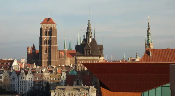 Widok na zabytkowe kamienice i wieże kościołów w śródmieściu Gdańska. W tle nowoczesna, miedziana bryła budynku.