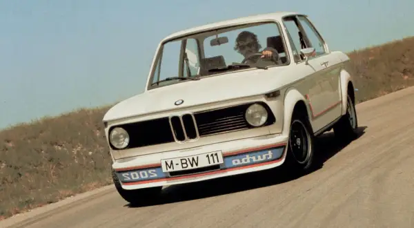 Białe BMW E20 z kolorowymi pasami na zderzaku i czerwonymi akcentami na karoserii jedzie po krętej drodze.