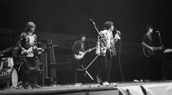 Vier Musiker auf einer Bühne, die alte Rockbands repräsentieren. Einer spielt Schlagzeug, die anderen Gitarre und Bass.