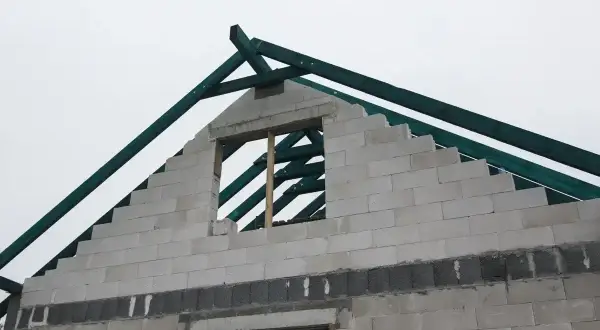 Budowa domu w toku. Widać szczytową ścianę z bloczków, na której powstaje wieniec, oraz zieloną więźbę dachową.
