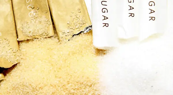 Zbliżenie na rozsypany cukier trzcinowy i biały, obok otwartych saszetek z napisem "SUGAR".
