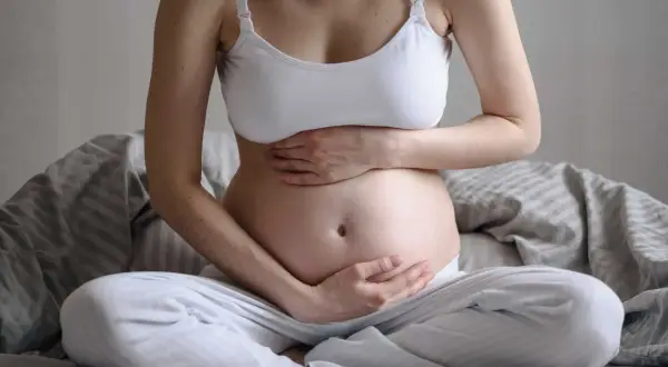 Gravid kvinna sitter på sängen med händerna på magen. Hon känner sig lite dålig i magen, en vanlig känsla under graviditeten.