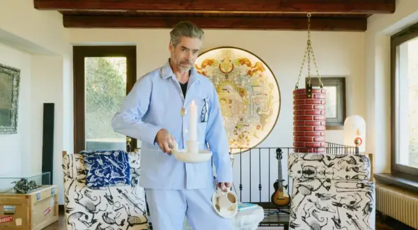 Uomo in pigiama azzurro tiene candela accesa e tazza di caffè, in un ambiente eclettico con opere d'arte e mobili bizzarri. Un'immagine che evoca un momento di riflessione, quasi un rituale mattutino, dove il lavoro incontra il sogno.