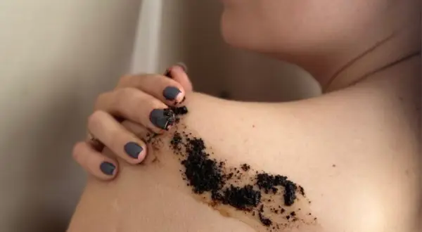 Dłoń z ciemnym lakierem do paznokci rozprowadza na ramieniu drobny, ciemny peeling z kawy.