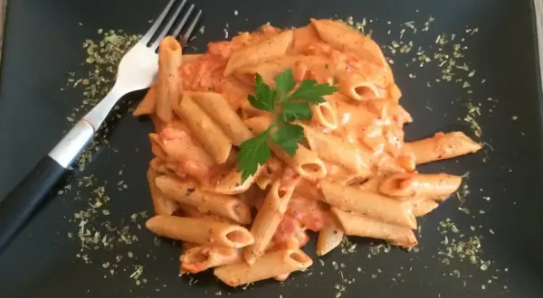 Penne w kremowym sosie pomidorowym z mascarpone, udekorowane natką pietruszki. Obok widelec i posypka z ziół.