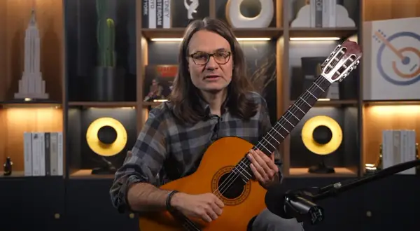 Mężczyzna z gitarą uczy, jak nauczyć się nut. W tle półki z książkami i dekoracjami.