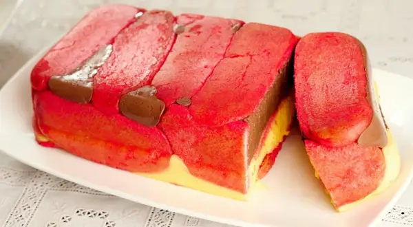 Una fetta di zuppa inglese tradizionale, con strati di pan di Spagna rosa, crema pasticcera gialla e cioccolato, decorata con savoiardi rossi e gocce di cioccolato.