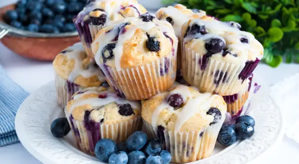 Stos puszystych muffinek z borówkami i białą czekoladą, polanych lukrem, ozdobiony świeżymi borówkami.