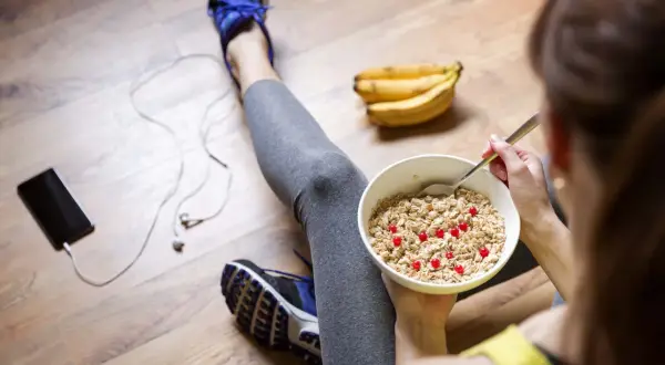 Après le sport, une femme mange un bol de flocons d'avoine avec des baies rouges. Elle est assise sur le sol en bois, à côté de ses chaussures de sport, d'un téléphone et d'écouteurs. Elle suit un régime pour maigrir.