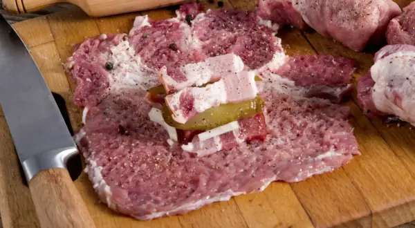 Przygotowanie zrazów z karkówki: rozbite plastry mięsa z boczkiem, ogórkiem i cebulą, obok przyprawy i nóż.