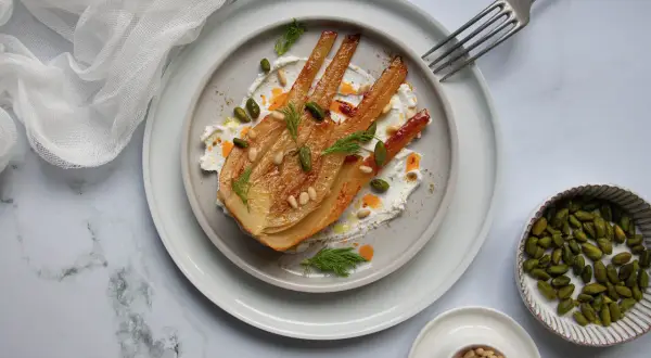 Un plat de fenouil au four, doré et tendre, servi sur une crème onctueuse, garni de pistaches, pignons et d'aneth frais.