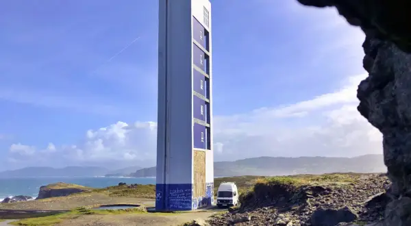 El faro de Punta Frouxeira se alza imponente sobre la costa, con un cielo azul y nubes blancas. Una furgoneta blanca descansa a su lado.