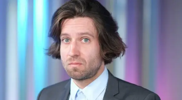 Michał Kempa stand up, w garniturze i jasnej koszuli, z burzą brązowych włosów i niebieskimi oczami, na tle kolorowych pasów.